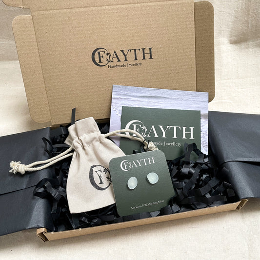 Pale blue sea glass stud earrings, on a branded dark green square display card are inside a brown cardboard box with black tissue paper, a branded beige cotton tie gift bag and a business card.