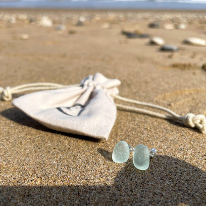 light blue sea glass studs on sand at the beach with a beige cotton tie gift bag in the background