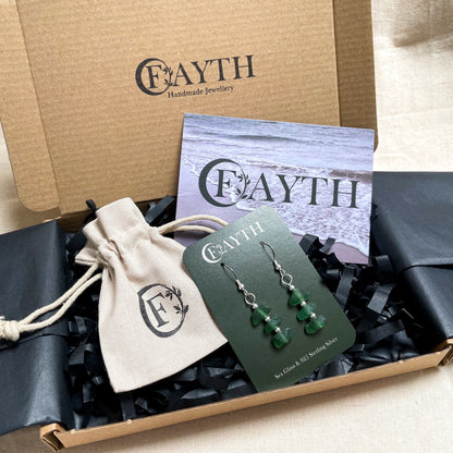 Green sea glass earrings on a green branded display card are inside a carboard box with black tissue paper, a branded beige cotton tie gift bag and a business card