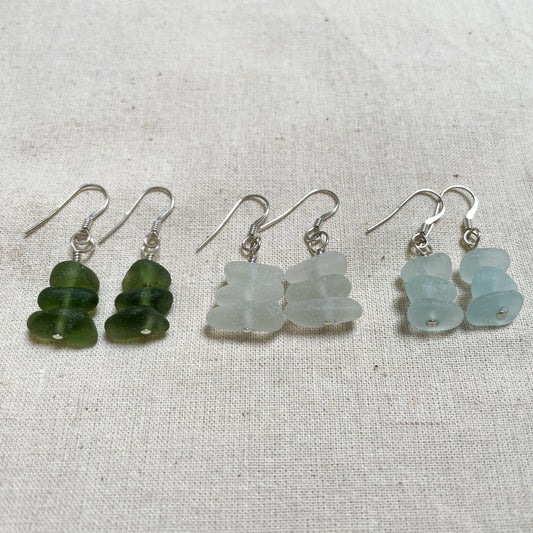 Three pairs of earrings laid on a beige linen background. Earrings are made up of three small pieces of sea glass connecting to a small silver ear wire. Green sea glass earrings (right), white sea glass earrings (middle), light blue sea glass earrings (left)