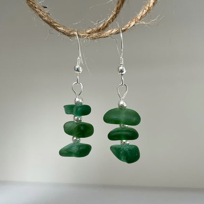 green sea glass earrings made up of three small pieces of green sea glass with three small silver beads in between, attached to a silver ear wire with the same silver bead. earrings are hanging from a piece of string