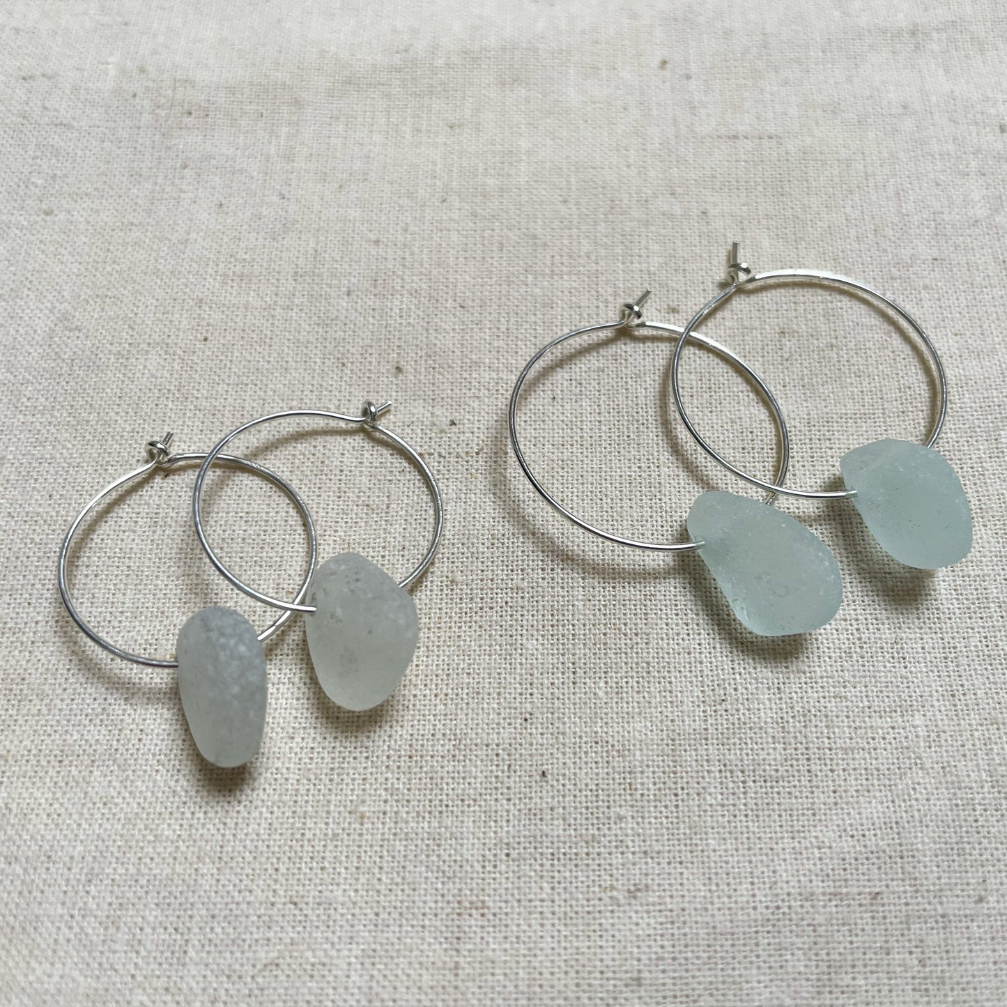 Two pairs of silver hoop earrings with sea glass on a beige background.One pair of hoops is smaller than the other, smaller white sea glass hoops (left), larger Light blue sea glass hoops (right)