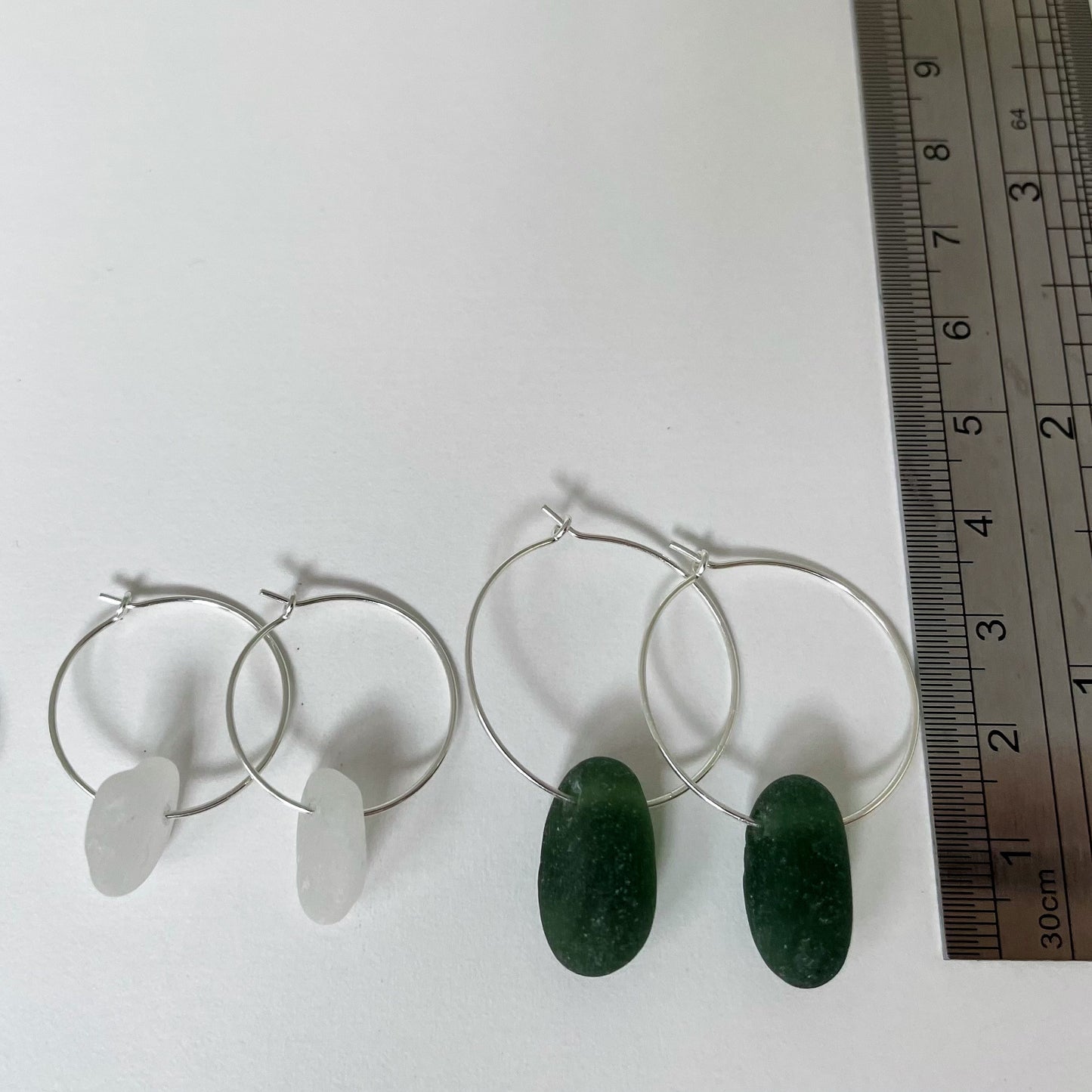 Smaller pair of white sea glass hoop earrings (left) next to a larger pair of dark green sea glass hoop earrings (right), are next to a ruler showing the height from the bottom of the sea glass to the top of the silver hoop on the larger dark green sea glass earrings around 3.5cm