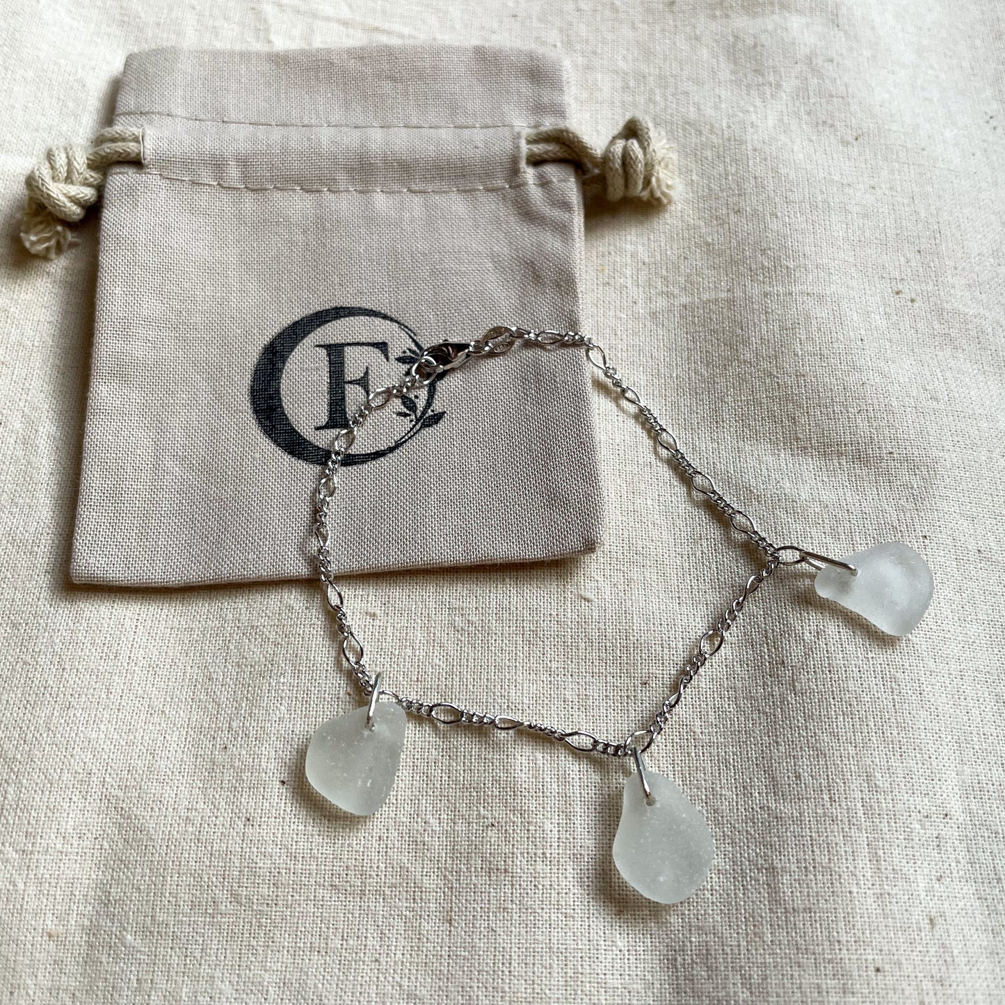 Sterling silver figaro chain bracelet with three pieces of white sea glass on branded beige cotton tie gift bag.