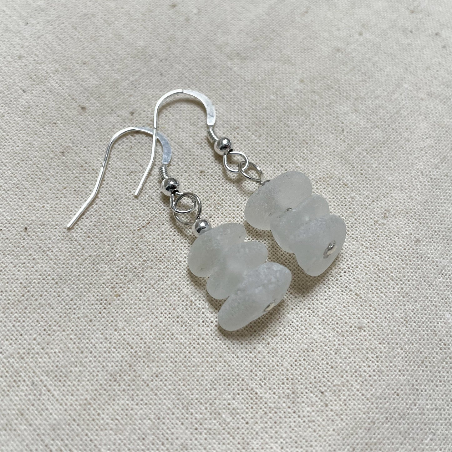 White sea glass dangle earrings made up of three pieces of small white sea glass with a silver bead on top, connecting to a Shepard's hook ear wire in silver with a silver bead on