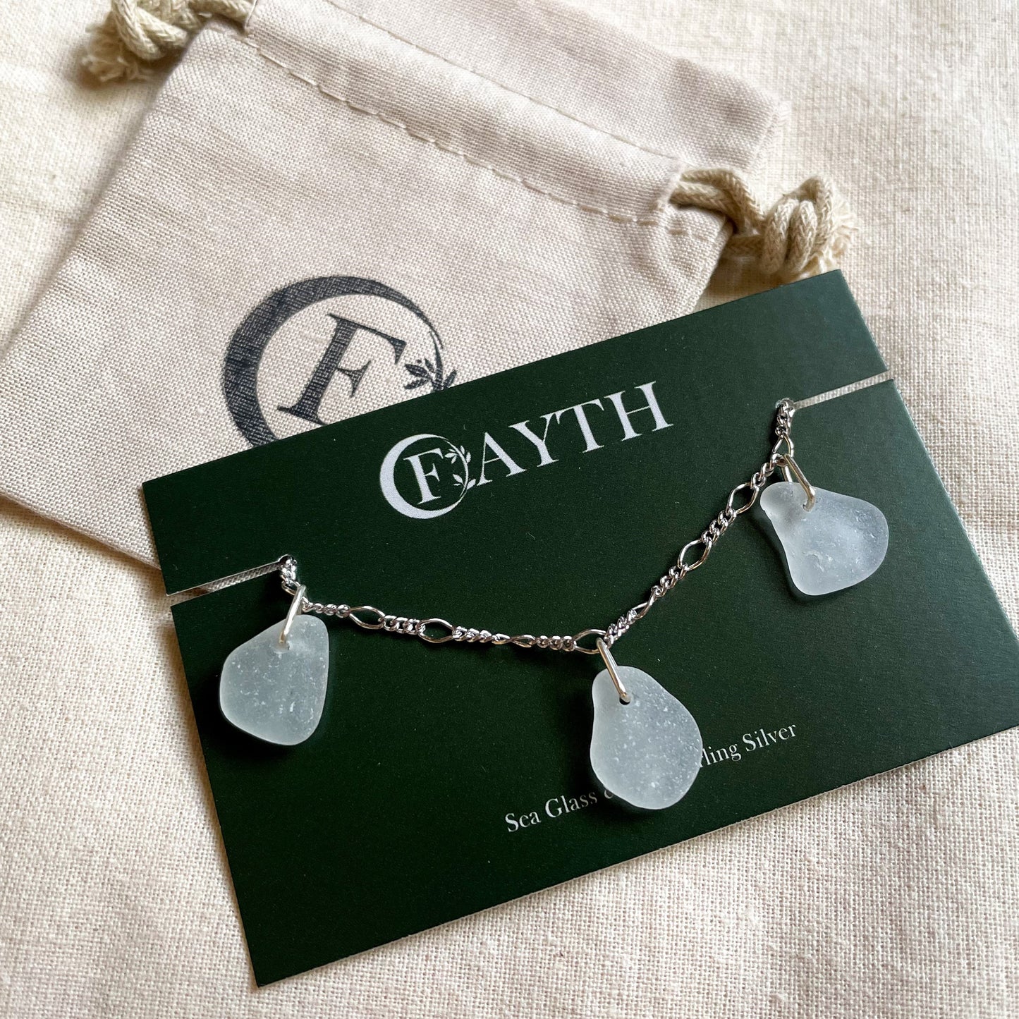 Sterling silver figaro chain bracelet with three pieces of white sea glass on a dark green branded display card, behind is a branded beige cotton tie gift bag.