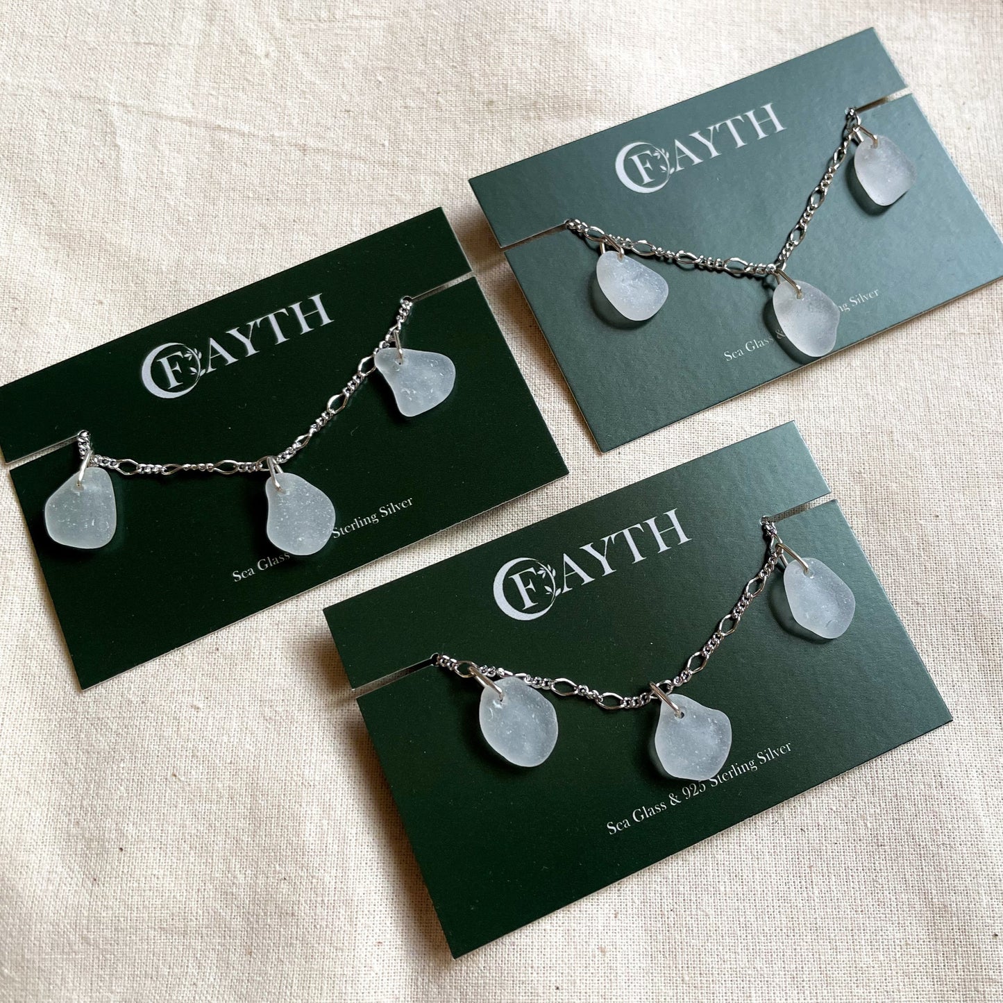 Three sterling silver white sea glass figaro bracelets on dark green branded display cards.