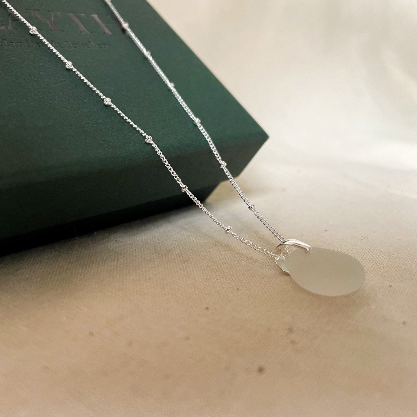 White sea glass necklace on silver satellite chain hanging over dark green gift box